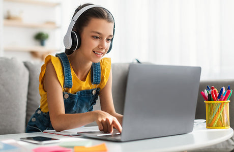 Mädchen sitzt am Schreibtisch und tippt auf der Tastatur ihres Laptops, trägt dabei Kopfhörer.